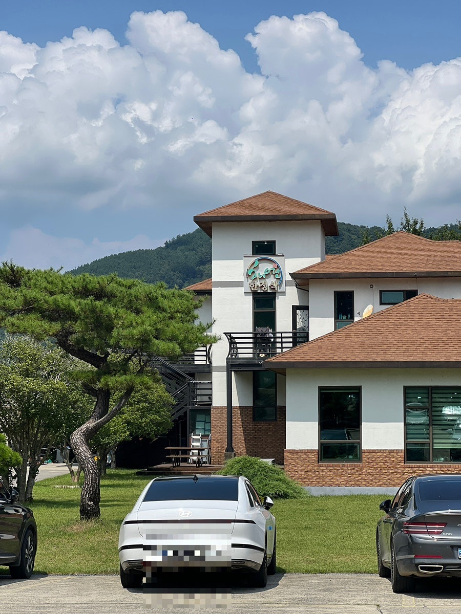 세련된 한옥 스타일의 건물 전경. 산을 배경으로 지어진 2층 건물이 넓은 잔디밭 위에 서 있고, 소나무와 계절 꽃이 어우러진다.