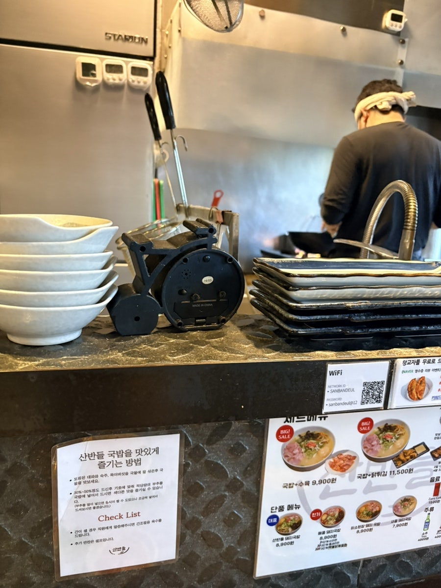 그릇을 정리하는 주방 모습. 카운터에는 와이파이 정보와 '국밥 맛있게 먹는 법' 안내문이 붙어 있습니다.