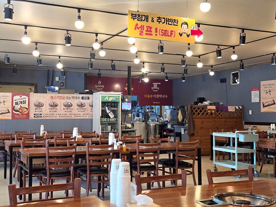 매장 내부 모습. 천장에는 '부침개&추가반찬 셀프!' 안내판이, 벽에는 메뉴와 가격표가 붙어 있습니다. 물막국수·비빔막국수·들기름막국수는 각 14,000원, 고단백막국수는 15,000원입니다.